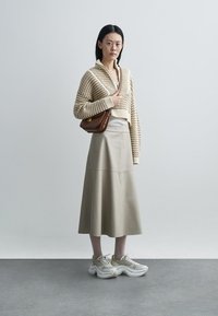 Kvinde stående iført beige strikket sweater, lang beige nederdel, chunky sneakers og holder en brun skuldertaske mod en ensfarvet hvid baggrund.