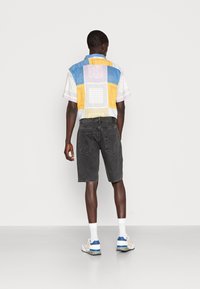 Camisa de manga corta con un patrón multicolor combinada con shorts de mezclilla negros, calcetines blancos y zapatillas con detalles azules, vista desde atrás.