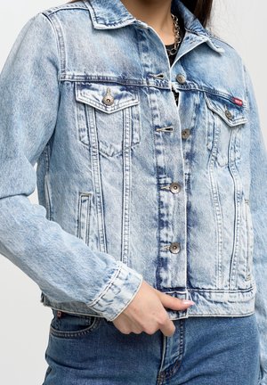 Persona que lleva una chaqueta vaquera de lavado ácido azul claro con botones de metal y bolsillos en el pecho, combinada con vaqueros de mezclilla azul oscuro.