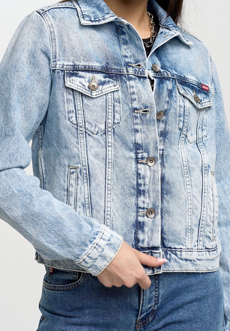 Personne portant une veste en denim bleu clair délavé à l'acide avec des boutons métalliques et des poches poitrine, associée à un jean en denim bleu foncé.