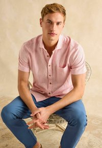 Heled roosa lühikeste varrukatega särk kergelt tekstuuriga, viis nuppu ja rinnatasku, millele on sobitatud sinised püksid ning istub korvtoolis.