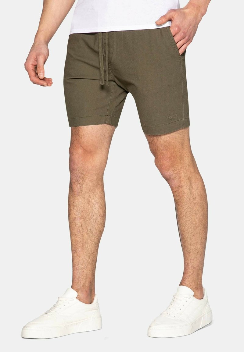 Homme portant un short vert olive avec cordon de serrage et des baskets blanches, debout avec une main dans la poche sur un fond uni.