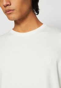 Hvid bomuldscrewneck sweatshirt med et diskret præget logo på venstre bryst, glat tekstur og klassisk pasform.