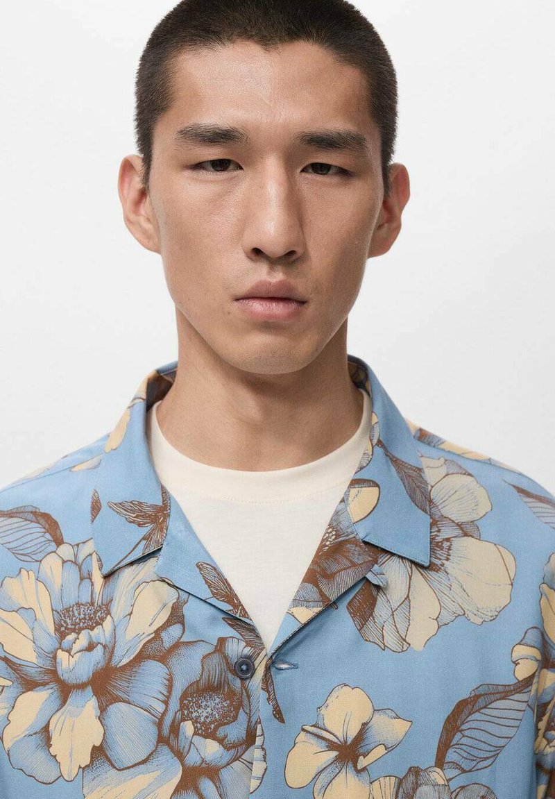 Kurzärmeliges Hemd in Hellblau mit einem Blumenmuster in Beige und Braun. Mit spitzem Kragen und einem einzelnen Knopf vorne.