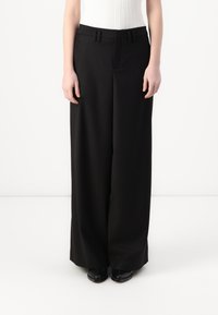 Pantalon noir à jambe large en tissu lisse, avec une taille plate, des passants de ceinture et des poches latérales. Associé à des chaussures noires.