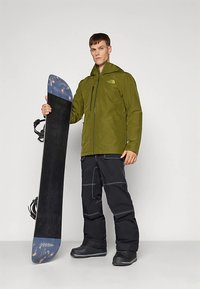 Giacca impermeabile verde con cerniera frontale, pantaloni da neve neri e snowboard nero con punta blu, tenuti da un modello maschile in piedi.