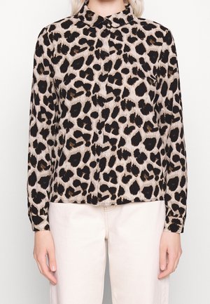 Bluzka na guziki z długim rękawem w czarno-brązowy wzór w cętki, zestawiona z beżowymi spodniami, noszona przez stojącą osobę.