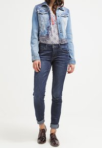 Kvinna med en ljusblå denimjacka, mönstrad blus, mörkblå uppkavlade jeans och blanka bruna snörskor stående med avslappnade händer.