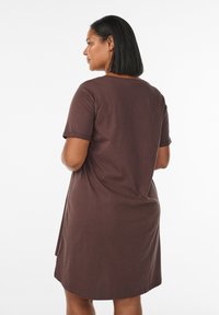 Donna con capelli neri di lunghezza media che indossa un vestito marrone a maniche corte lungo fino al ginocchio, in piedi di spalle su uno sfondo bianco.