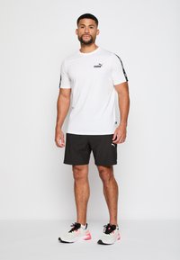 Camiseta blanca de manga corta con el logo negro de Puma en el pecho, combinada con pantalones cortos negros y zapatillas deportivas de colores variados con acentos negros.