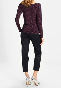 Mujer con una blusa oscura de manga larga con un patrón de corazones rosas, pantalones negros texturizados y zapatos de tacón alto plateados, de pie de espaldas.