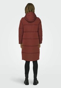 Manteau long rembourré rouge avec capuche, mettant en avant des sections matelassées horizontales. Porté avec un legging noir et des bottes noires épaisses.