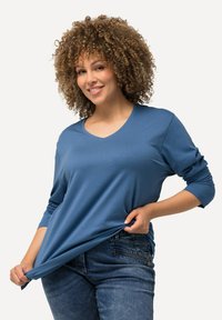 Blaues Langarm-V-Ausschnitt-Shirt aus einem weichen Stoff. Der Träger hält das Shirt, während er eine blaue Jeans trägt. Der Hintergrund ist schlicht.