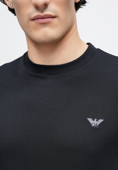 Juoda medvilninė marškinėliai su užapvalinta iškirpte ir mažu siuvinėtu erelio logotipu kairėje krūtinės pusėje. Lygus paviršius, minimalistinis dizainas.