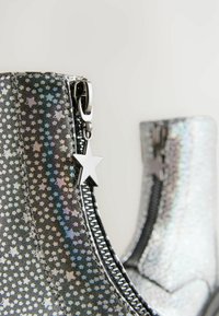 Srebrne botki do kostki z gwiazdkowym wzorem, błyszczący materiał, czarny zamek błyskawiczny i uchwyt w kształcie gwiazdy. Gładka tekstura i elegancki design.