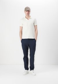 Bílý žebrovaný polo tričko s V-výstřihem, spárované s námořnickými kalhotami. Doplňuje bílé tenisky a béžovou kšiltovku. Model stojí s rukama v kapsách.
