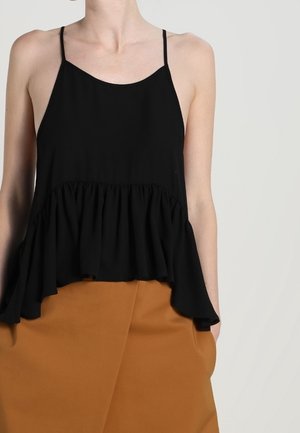 Mujer con top negro sin mangas con volantes y tirantes finos y falda marrón de talle alto sobre un fondo claro y liso.