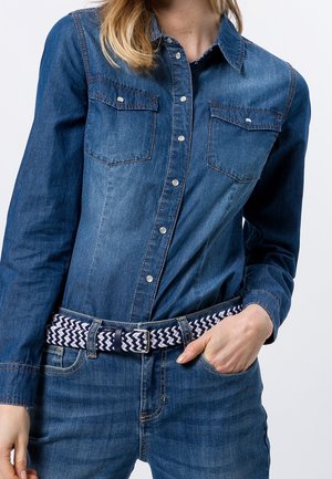 Frau trägt langärmliges Jeanshemd mit Druckknöpfen und zwei Brusttaschen, kombiniert mit blauen Jeans und marineweißem gewebtem Gürtel.