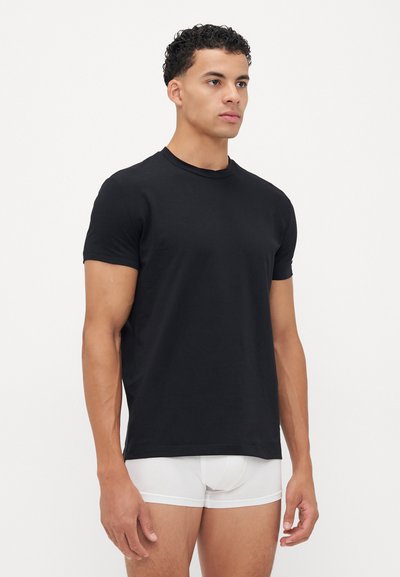 Mann steht entspannt mit den Händen, trägt ein einfaches schwarzes T-Shirt und weiße Boxer-Shorts vor einem schlichten, hellen Hintergrund.