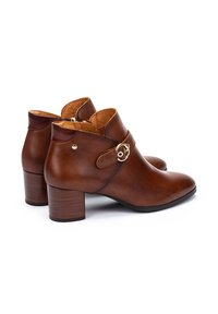 Botas de cuero marrón hasta el tobillo con punta redonda, tacón medio y una banda decorativa con un detalle de hebilla dorada. Textura suave con acentos de costura.