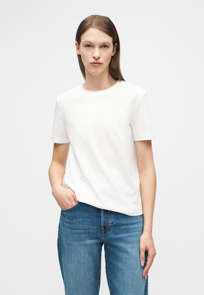 Νεαρός με ίσια καστανά μαλλιά φοράει απλό λευκό κοντομάνικο μπλουζάκι και μπλε τζιν, στέκεται με το ένα χέρι στην τσέπη μπροστά σε λευκό φόντο.