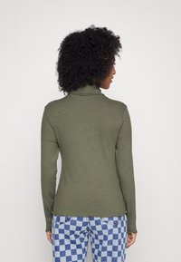 Sweater de gola alta canelado em verde-oliva com mangas longas, design ajustado e um detalhe sutil na bainha, combinado com calças xadrez azuis.