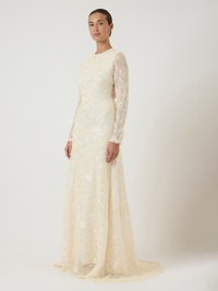 Γυναίκα με μακριά μανίκια, σε φόρεμα από κρέμα δαντέλα, μήκους μέχρι το πάτωμα και με λουλούδια, που στέκεται μπροστά σε απλό ανοιχτό φόντο.