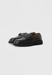Black leather loafers with textured surface, metal buckle detail, and thick, ridged black sole. Classic design with a low profile.