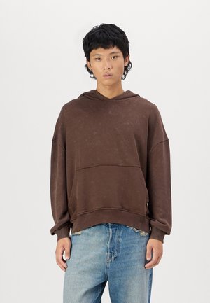 Brun overdimensjonert hettegenser med tekstur, frontlomme og snorhette, parret med lyseblå slitte jeans.
