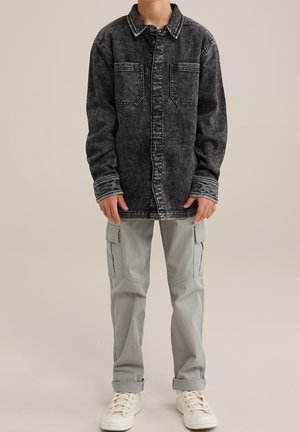 Barn iført sort acid-vasket denimjakke, lysegrå cargobukser og hvide snøresneakers, stående mod en ensfarvet baggrund.