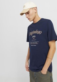 Tums navy zila izgatavota no kokvilnas ar drukātu logotipu gaišā brūnā krāsā, apvienota ar bēša tintinieka cepuri, modele tur kabatu džinsu šortos.