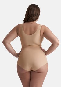 Plus-size kvinna med långt brunt hår som bär beige sömlöst shapewear bakifrån, händerna vilande på höfterna mot en neutral bakgrund.