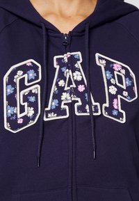 Felpa con cerniera di colore blu navy, con logo "GAP" a motivo floreale in bianco e fiori multicolori. Presenta lacci e una morbida texture in pile.