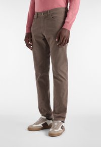 Mann in braun karierten Hosen, weißen und braunen Turnschuhen und einem pinken Langarmshirt, steht vor einem weißen Hintergrund.