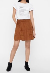 Weißes T-Shirt mit schwarzem Druck, kombiniert mit einem gestuften braunen Wildlederrock. Schwarze knöchelhohe Stiefel mit dekorativen Nieten.