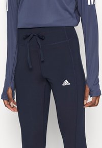 Granatowe legginsy sportowe z dopasowanym krojem, z elastycznym paskiem w talii oraz białym logo Adidasa blisko lewej biodra. Gładki, rozciągliwy materiał.