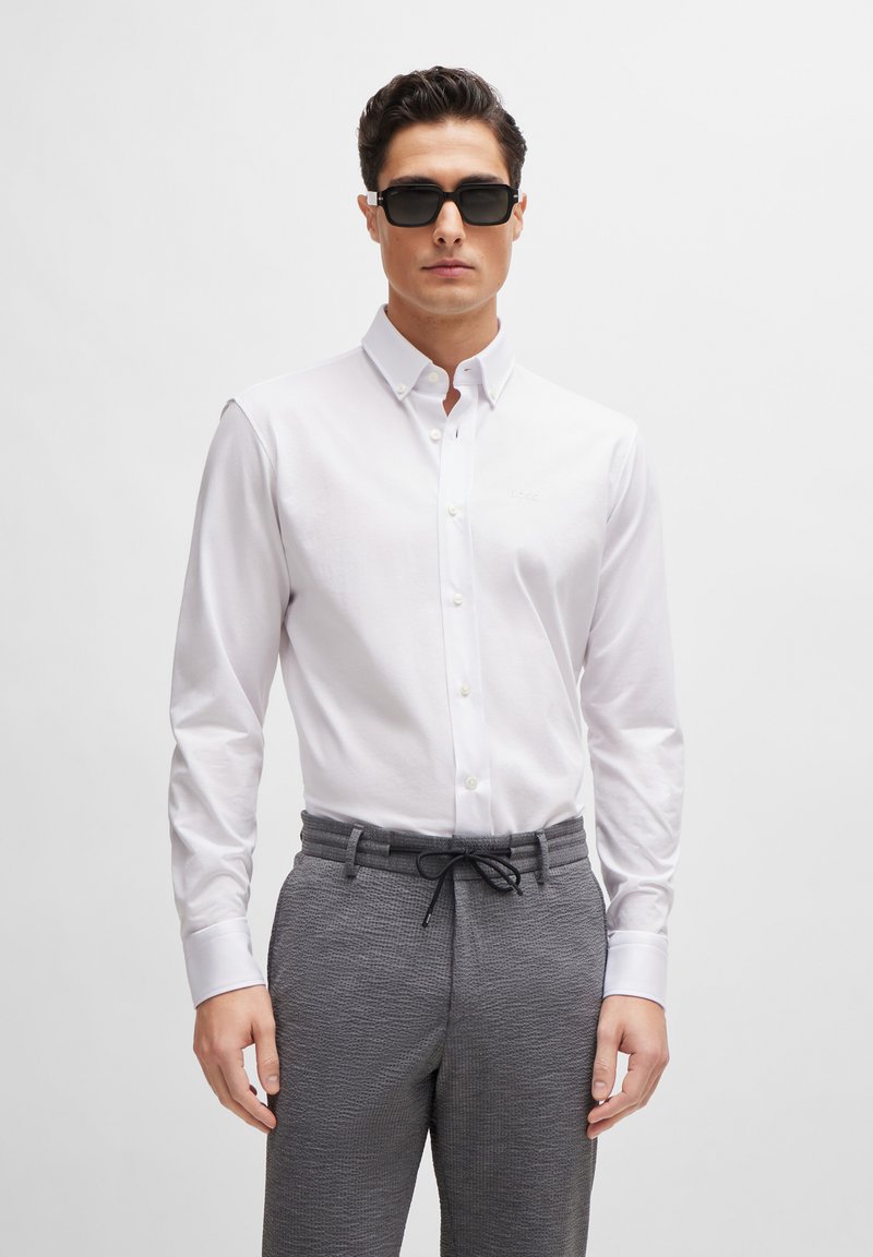 Vit button-down skjorta med krage; grå texturerade byxor med dragsko i midjan; modell med svarta solglasögon.