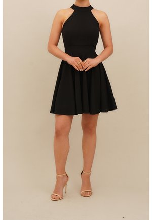 Mujer que lleva un vestido negro sin mangas con cuello halter y sandalias beige de tacón alto, de pie con las manos entrelazadas delante.