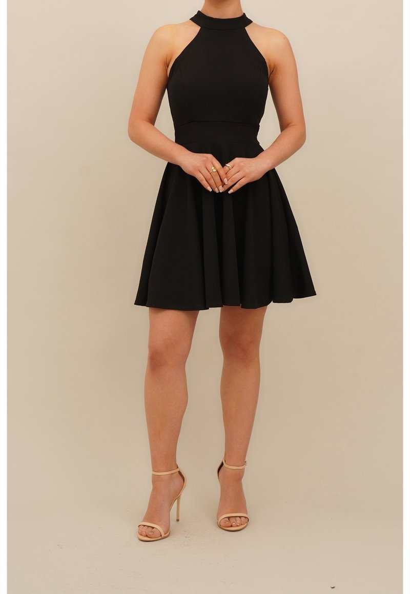 Femme portant une robe noire sans manches à col halter et des sandales beige à talons hauts, debout, les mains jointes devant.