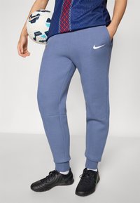 Lyseblå Nike sweatpants med skrå lommer, ribbestrikkede mansjetter og en jevn tekstur. En svart fotball med et geometrisk mønster holdes.