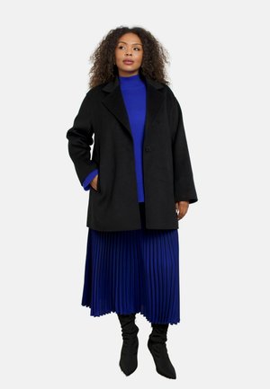Zwarte wolle jas met een notch-kraag, gedragen over een blauwe coltrui en een geplooide blauwe rok. Kenmerkt zich door kniehoge zwarte laarzen.