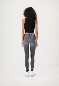 Femme aux cheveux bouclés portant un crop top noir sans manches, un jean skinny gris et des baskets blanches montantes, debout, de dos.