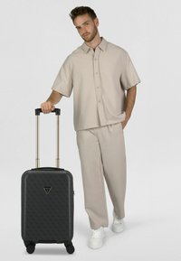 Hombre con atuendo beige de pie, con una mano en el bolsillo y la otra sujetando el asa de una maleta negra con diseño.