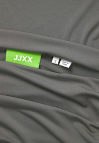Sivi materijal s glatkom teksturom, sadrži zeleni etiketu s natpisom "JJXX" i oznaku veličine "S" pored etikete "MADE IN CHINA".