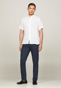 Camicia bianca in lino a maniche corte con bottone frontale, abbinata a pantaloni blu navy e scarpe nere, con un piccolo logo sulla camicia.