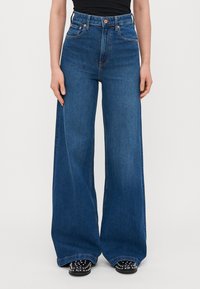 Blå högmidjade vida jeans i denim med en ren design, fem fickor och kontrasterande orange sömmar. Platt fåll vid anklarna.
