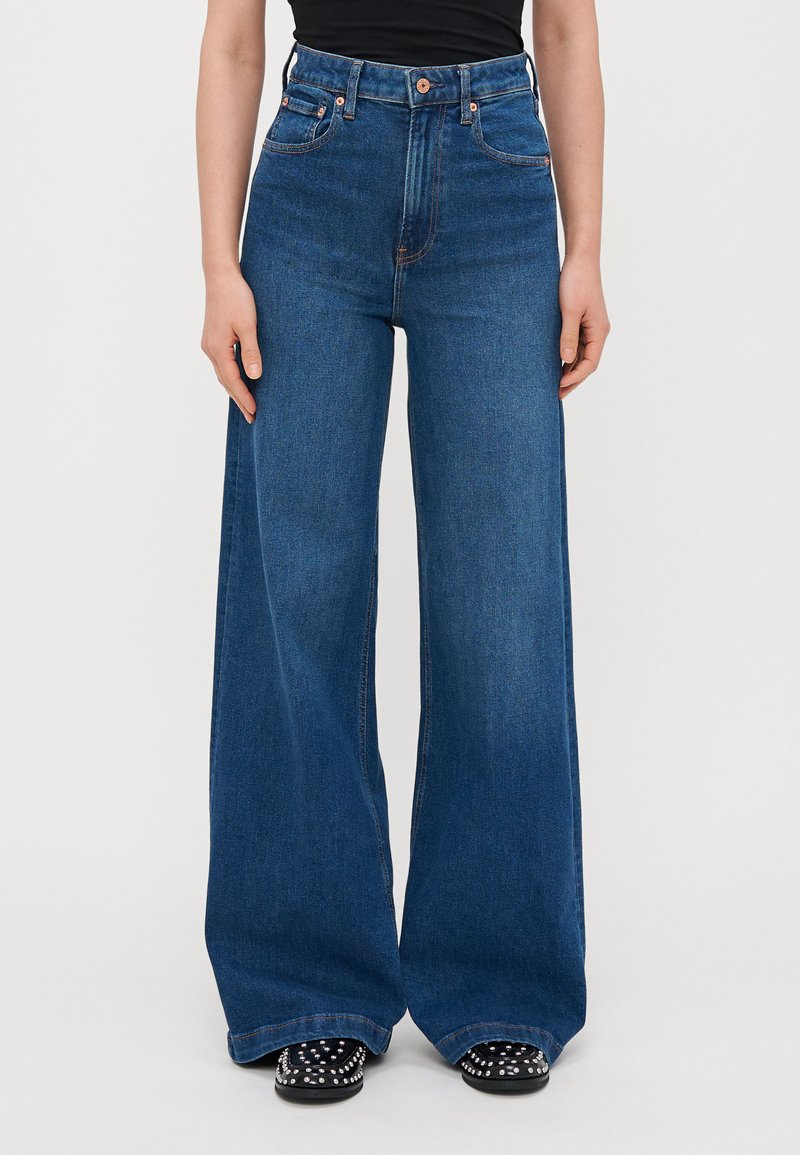 Blå högmidjade vida jeans i denim med en ren design, fem fickor och kontrasterande orange sömmar. Platt fåll vid anklarna.