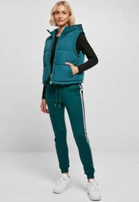 Gilet rembourré turquoise avec capuche et fermeture éclair, associé à un pantalon vert avec des rayures blanches sur les côtés. Le modèle porte des baskets blanches.
