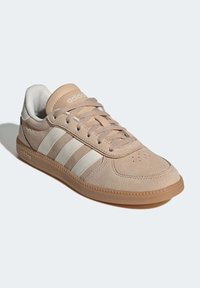 Beige atletisk sneaker med tre vita ränder, överdel i mocka, rundad tå och gummisula. Har snörning och vadderad krage.