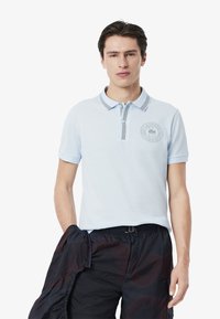 Ljusblå polo med randig krage och broderad logotyp på bröstet. Kortärmade med en slät textur, kombinerat med mörka shorts.